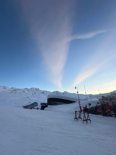 The image displays a snow-covered mountain ski resort in Saint-Martin-de-Belleville, France, under a clear blue sky with distinct cloud formations. The foreground is dominated by a smooth, white expanse of packed snow, indicative of a ski slope. In the midground, from left to right, a small, white building with a grey pitched roof is partially buried in snow. Adjacent to it stands a large, modern, multi-level building featuring a curved dark facade with vertical elements and expansive glass sections, marked with the visible signage "ALAYSA." Further to the right, another multi-story building with multiple red outdoor umbrellas or awnings is visible, suggesting a restaurant or commercial establishment.
In the mid-right foreground, two dark wooden A-frame structures rest on the snow. A tall black lamppost with a curved arm and lantern-style fixture is positioned to the right of the "ALAYSA" building, alongside several red and white striped ski marker poles and a small evergreen tree. The background consists of rugged, snow-capped mountain peaks under a vibrant blue sky. Two prominent, wispy white cloud formations ascend from the lower mid-left and mid-right, converging towards the upper center of the frame, creating a V-shaped or fan-like appearance. The lighting suggests either early morning or late afternoon, casting an even, cool illumination across the scene. No individuals are visible.