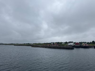 The image depicts a coastal scene in Sleneset, Norway, under a cloudy sky. The foreground is dominated by dark, choppy water. In the middle ground, a rocky shoreline extends from the left, forming a breakwater. A small settlement of houses, primarily red with white trim and some white buildings, is situated along the elevated land behind the breakwater. Trees are visible in the background. The houses appear to be traditional Norwegian coastal homes. The overall atmosphere is overcast and subdued.