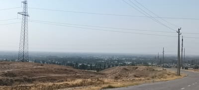 The image shows a panoramic view of a semi-arid landscape with a city in the background. In the foreground, there is a dirt road or path winding through dry, brown vegetation on a hillside. To the left, a tall, lattice-like metal pylon stands prominently against the sky, supporting overhead power lines. The power lines stretch across the frame, disappearing into the distance.

Further down the hill and extending into the middle ground, a paved road curves to the right. Along this road, several utility poles with power lines are visible. Beyond the road and the immediate hillside, a sprawling city is visible in the hazy distance. The city is characterized by a dense collection of buildings and trees, suggesting an urban or semi-urban area. The overall atmosphere is hazy, likely due to dust or atmospheric conditions, obscuring distant details. The sky is a pale, washed-out blue. The scene appears to be a view from an elevated position overlooking the city of Margilan, Uzbekistan.