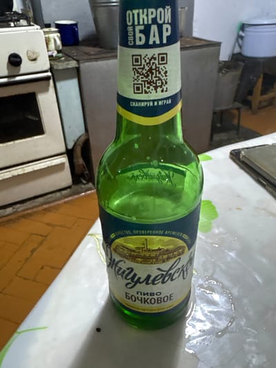 A green glass beer bottle labeled "ЖИГУЛЕВСКОЕ ПИВО БОЧКОВОЕ" and "ОТКРОЙ СВОЙ БАР" stands on a white, slightly wet surface. The bottle has a QR code printed on its label. In the background, a white stove and a blue mug are visible. The floor is made of orange tiles. The overall scene appears to be inside a kitchen.