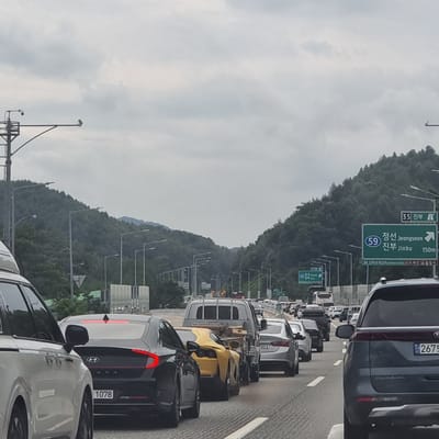 This image captures a traffic jam on a highway in Jinbu-myeon, South Korea. Multiple lanes of vehicles are stationary or moving slowly. The sky is overcast, and the surrounding environment consists of dense, green, tree-covered hills.

Prominent vehicles in the foreground include a white SUV on the left and a dark grey Kia Carnival SUV on the right, identifiable by its license plate which reads "2675". Behind the white SUV is a black Hyundai Sonata with a license plate "1078". A bright yellow sports car is visible in front of a silver sedan. A small truck with a trailer is also in the traffic flow.

Road signs are visible, including green directional signs. One sign indicates "35 Jinbu" and another indicates "59 Jeongseon, Jinbu" with an arrow pointing right, along with a distance of "150m". Streetlights and traffic cameras are mounted on poles along the highway. The road is paved, with visible lane markings. The overall scene depicts a typical highway congestion in a rural or semi-rural mountainous region of South Korea.