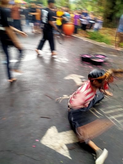The image depicts a blurred, dynamic outdoor scene on a wet, dark asphalt road in Mojogedang, Indonesia.
In the foreground, an individual is captured in a low, crouching or falling posture, facing left. This person wears a dark head covering, a red and white vertically striped top, and dark pants. Long, light brown hair is visible, flailing with motion. A rope or twine is visibly secured to the lower back area of this individual, extending to the left. A light-colored shoe is visible on the right foot.
In the midground, two black, open containers or bowls are on the asphalt, each holding a bright pink substance, which is also scattered in fragments across the wet road surface.
To the left of the frame, two barefoot individuals are in motion, moving from left to right. The closest figure wears dark clothing, with the right arm extended forward and the left leg raised in a bent, striding position. The second figure, slightly behind, wears a black t-shirt with a white graphic on the back and carries a dark, thin stick or whip in their right hand.
The background shows a blurred crowd of people in various colored attire, suggesting spectators or participants. An orange cylindrical object, possibly a drum or large pot, is also visible. Green foliage lines the background, indicating an outdoor setting. A large, white directional arrow is painted on the asphalt surface, pointing left, located beneath the crouching figure.