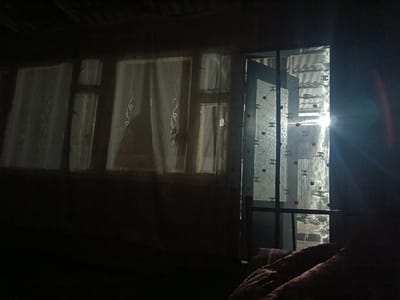 The image depicts a dimly lit interior with natural light emanating from a window and a glass door on the right side. The light source appears to be intense, creating lens flare and washing out details outside. Inside, white sheer curtains hang across multiple windows on the left, with subtle decorative patterns. A glass door on the right is adorned with a repeating pattern of interlocking "C" symbols and small red circular elements. The overall scene suggests a residential setting, possibly a bedroom, with a dark, textured blanket or bedding visible in the foreground on the right. The environment outside the windows and door appears to be constructed of corrugated metal or similar material, indicative of a functional or possibly rustic architectural style. The location is identified as Sretenka, Kyrgyzstan.