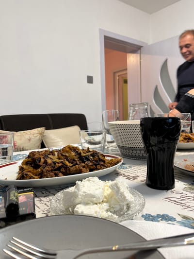 A table is set for a meal with food and beverages. Dishes on the table include caramelized onions, fresh white cheese or cream cheese, and what appears to be a Coca-Cola bottle. Cutlery and plates are present, suggesting a meal in progress or about to begin. A man is visible partially in the background, out of focus. The tablecloth has a floral design and text, which is illegible. The location is not discernable from the image. Karavelovo, Bulgaria cannot be determined.