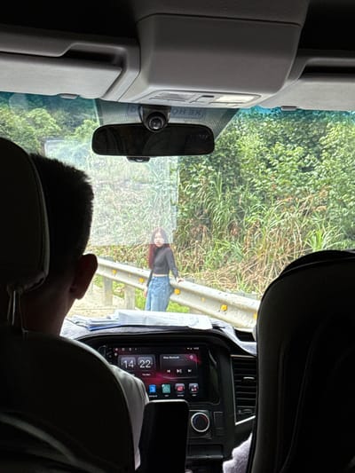 The image, captured from inside a vehicle, shows a young woman with long, reddish-brown hair standing on the side of a road. She is wearing a black long-sleeved crop top and blue denim jeans. She is leaning on a yellow guardrail that separates the road from a densely vegetated area. The greenery consists of tall grasses and trees, suggesting a rural or semi-rural environment, possibly located in the city of Nam Khanh Vinh, Vietnam. Through the vehicle's windshield and reflected on its surface, the woman is visible. The interior of the vehicle is also visible, including the back of a person's head in the foreground, a rearview mirror, and the dashboard with a digital display showing the time as 14:22.