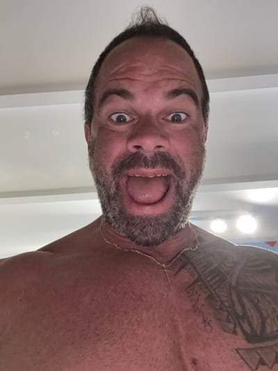 A close-up, rotated portrait captures a man looking directly at the camera with a wide-open mouth and wide eyes, displaying an exaggerated, perhaps playful, expression. The image is rotated 90 degrees counter-clockwise. The man has dark hair with some graying, a full beard and mustache, and visible forehead wrinkles. He appears shirtless, revealing a prominent dark tattoo with tribal or abstract patterns on his right shoulder and upper arm. A thin, gold-colored chain with a small pendant (possibly a cross) is visible around his neck. The skin on his chest and shoulder has a reddish tone. The background consists of a plain, light-colored ceiling or wall, with two bright, circular light sources in the upper right suggesting an indoor setting with artificial lighting. No text is visible. The scene takes place indoors in Bredene, Belgium.