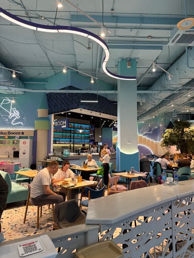 The image depicts the interior of a cafe called "Boom Cafe". The cafe is located in Перовское сельское поселение, Ukraine.
Several people are present. Two men are seated at a table, appearing to be in conversation. Other individuals are seated at tables in the background, suggesting a cafe environment.
The interior design is modern with a color palette of light blues and purples. The ceiling is exposed, revealing pipes and wires. The cafe features distinctive elements, including a column resembling a tower and a wall decorated with a cartoonish depiction of a rocket. The cafe's name, "Boom Cafe", is prominently displayed.
The cafe is populated with tables and chairs, and details such as the patterned floor and decorative wall designs contribute to the overall ambiance of a contemporary dining space.