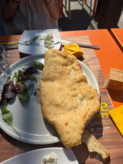 The image captures a culinary scene on an orange table in Pompei, Italy. A fried bread rests on a white plate with a cartoon character design. A side of mixed greens accompanies the bread. A piece of the bread is broken off. Adjacent to the plate is a smaller dish containing a creamy white substance, potentially cheese. Utensils, including a fork and knife, are present near the cheese dish. A wooden block with the number "47" is visible, along with a yellow napkin and a leaflet displaying information related to "LE DELIZIE POMPEI," including their social media handles and website. A partially visible drinking glass sits at the edge of the frame. A person's arm and clothing are seen on the left. The setting suggests an outdoor dining experience.