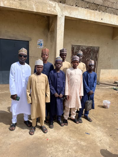 A group of seven people stand together in Naraguta Hausawa, Nigeria. They wear clothing typical of Hausa culture, including embroidered caps. The person at the front is wearing sunglasses and holding a phone. They stand in front of a weathered building. A political poster is partially visible on the wall. A plastic container rests on the ground near the group.