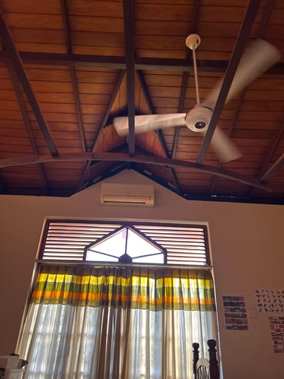 The image depicts an indoor scene, likely a room, with a wooden vaulted ceiling supported by dark beams. A white ceiling fan with its blades in motion is visible against the ceiling. Below the ceiling, an air conditioning unit is mounted on the wall. A large window with louvred shutters and a triangular pane at the top is covered by curtains. The curtains consist of a top section with horizontal stripes in yellow, green, and orange, and a bottom section of sheer, light-colored fabric. On the wall to the right of the window, there are several framed pictures or posters. The overall impression is of a warm, somewhat rustic interior. The location is identified as Dehiwala, Sri Lanka.