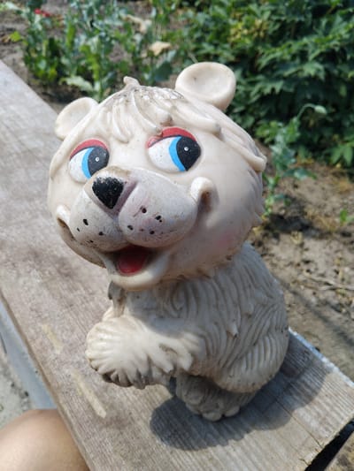 This media file shows a weathered, cream-colored toy animal, possibly a bear or a lion cub, lying on a wooden surface. The toy has large, round eyes painted with black pupils, blue and red accents, and white highlights. Its snout is painted black, with red accents on its cheeks and ears. The toy appears to be made of plastic and is showing signs of age, with some dirt and wear. Its fur texture is depicted with raised lines on its head and body.
The toy is positioned on a weathered wooden plank, which is placed outdoors. In the background, there is out-of-focus greenery, suggesting an outdoor setting like a garden or yard. The lighting suggests it is daytime, with a shadow cast by the toy onto the wooden surface, indicating a sunny day. The location is identified as Bila Krynytsia, Ukraine. No people are visible in the media file, and no specific activity or event is taking place; the toy is simply resting. There is no visible text in the image.