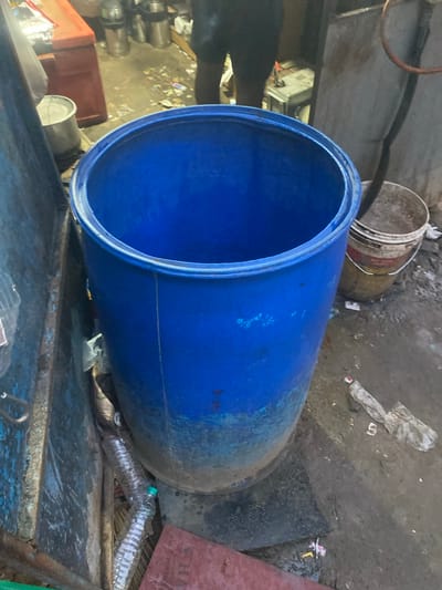 A blue plastic drum lies horizontally on a metal surface, its opening facing the viewer. The drum is large and appears to be empty. To the left of the drum, stacked metal cylinders are visible. In the background, a dirty, weathered bucket with a wire handle rests on the ground amidst debris. The ground is covered in dirt, scattered trash, and what appears to be mud. A red container is partially visible to the lower left of the blue drum. Two plastic water bottles are placed near the right side of the blue drum. The scene suggests an outdoor or industrial setting with elements of disarray and neglect. No specific time of day or weather conditions are discernible. There is no visible text.