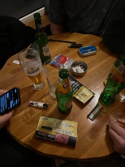 A wooden table is littered with beer bottles, a mostly empty glass, cigarette packs ("Camel," "Tobacco"), rolling papers, a lighter, a tin, and an ashtray filled with cigarette butts. A hand holds a smartphone with the screen illuminated, suggesting someone is using it. Other body parts, like a hand and clothing, are visible at the table's edges, indicating people are present. The overall scene suggests a casual social gathering involving drinking and smoking.