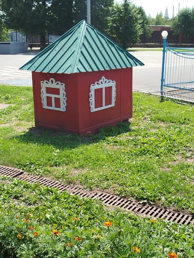 A small, red, dollhouse-like structure with a green, corrugated metal roof stands on a patch of grass. The structure has two white, decorative window frames on its visible side, each containing a simple white crossbar. In the foreground, there are orange marigold flowers and a drainage grate running horizontally across the image. The background shows a paved area, trees, a fence, a lamppost with a white globe, and a distant church spire. This scene is located in Yalutorovsk, Russia.