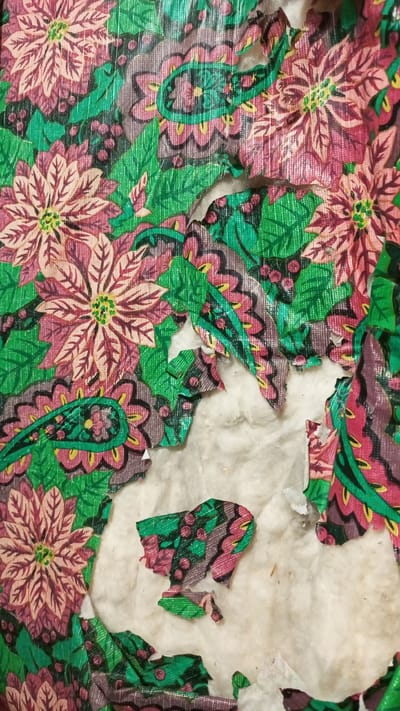 A close-up shot shows damage to patterned wallpaper. The wallpaper features green leaves, pink and maroon poinsettias, and paisley-like designs. Sections of the wallpaper are torn away, revealing underlying white insulation material. The image suggests deterioration or removal of the wallpaper. There is no information to indicate that this image was taken in Юдино, Russia.