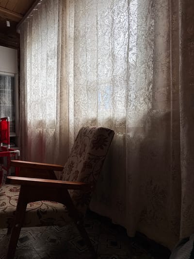 An interior scene with a wooden chair in the foreground and sheer curtains with a floral pattern covering a window in the background. The lighting suggests it is daytime, with light filtering through the curtains. The chair has wooden armrests and a patterned fabric seat and back. The floor has a geometric tile pattern. The environment appears to be a domestic setting.