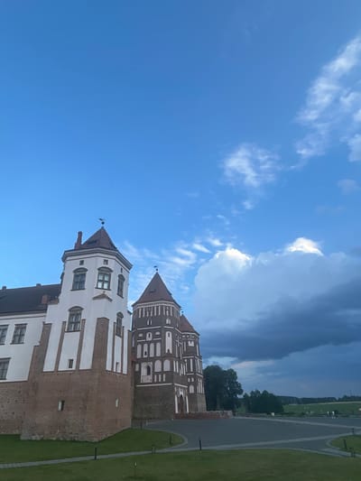The image captures a view of the Mir Castle Complex, a UNESCO World Heritage site located in the vicinity of Zhukhavichy rural council, Belarus. In the foreground, manicured green grass slopes gently upwards towards the castle. A paved pathway curves through the grass, marked by a few small, upright black bollards and some embedded lights.
The castle itself dominates the left and central portions of the frame. The left section features a tall, white, square tower with a reddish-brown tiled roof topped with a weather vane. This tower has multiple windows, some with dark frames, and is constructed with a base of rough-hewn reddish-brown stone and brick. Adjacent to it, to the right, is a circular tower with a conical, reddish-brown tiled roof, also featuring a weather vane. This tower is distinguished by its intricate brickwork in a pattern of alternating dark red and lighter shades, with numerous arched windows and decorative white elements. The roofs of both sections are steep and tiled.
The sky above is a vibrant blue, with fluffy white clouds scattered across the upper left and a large, dark, ominous cloud formation gathering in the upper right, suggesting an approaching storm. A few distant, dark green trees are visible behind the castle, bordering a field. The overall impression is of a historic architectural landmark set against a dynamic sky.