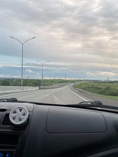 View from inside a car driving on a multi-lane highway, likely on a bridge or overpass, in Bol'shaya Mos', Russia. The road is straight with light traffic. There are lampposts along the side and fields with power lines beyond the highway. The sky is overcast. The dashboard of the car, including a circular vent with a five-pointed design, is visible in the foreground.