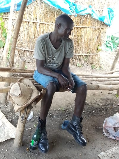 A man, appearing to be of Nigerian descent, is seated on a rustic wooden bench in Otulu, Nigeria. He is wearing a grey t-shirt with a graphic design and blue denim shorts. His legs are crossed, and he holds a lit cigarette in his right hand. On the bench beside him rests a tan leather hat with decorative stitching. A green glass bottle lies on the ground near his feet, along with a black leather boot. Another black boot is on his left foot, and a blue sandal is partially visible on the ground to his left. The background consists of a wall constructed from dried reeds and a blue tarp stretched overhead, suggesting an outdoor or semi-enclosed structure. The ground is composed of dirt and small stones.