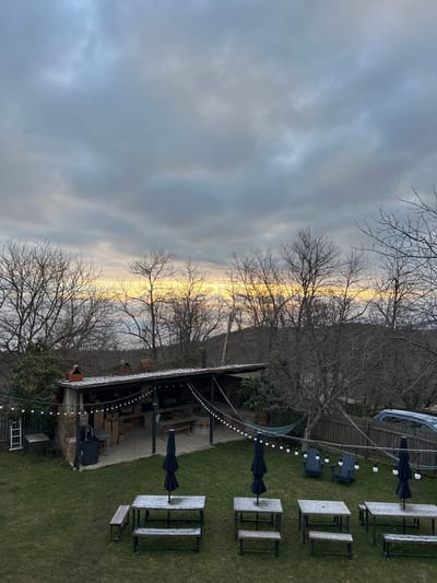 The image depicts an outdoor recreational area, likely a guesthouse or restaurant setting, in Sighnaghi, Georgia, under an overcast sky during what appears to be a cold period, possibly early morning or late afternoon given the light. The upper third of the frame is dominated by heavy grey clouds, with a distinct, bright yellowish-orange band of light visible at the horizon line, suggesting either sunrise or sunset. Bare deciduous trees, characteristic of late autumn or winter, populate the background and midground, with some evergreen foliage visible near structures.
In the midground, a large, rectangular, open-sided covered structure with a dark, snow-dusted sloped roof is present. This structure features brick walls, multiple wooden tables and benches within, and an integrated brick oven or fireplace on its left side. String lights with white bulbs illuminate the interior of this pavilion. Adjacent to the structure, a white step ladder leans against a smaller shed on the far left. A blue hammock is strung between the main covered structure and a tree to its right.
The foreground consists of a green lawn lightly dusted with snow or frost. Four wooden picnic tables, each equipped with attached benches and a folded black patio umbrella, are symmetrically arranged on the grass. These tables also exhibit a light covering of snow. A string of white bulb lights extends diagonally across the lawn from the covered structure towards the right. Two dark-colored Adirondack chairs are positioned on the grass to the right, near a wooden fence. A light-colored vehicle is partially visible behind this fence on the right side of the frame. No individuals are present in the scene.