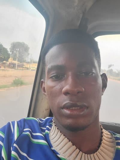 A close-up, selfie-style shot captures a young Black man inside a vehicle, possibly a bus or van, in what appears to be an overcast or dusty environment. The man is positioned centrally in the frame, with his face filling the majority of the image. He has short, dark hair, dark eyes, and medium-toned skin that appears slightly moist, suggesting perspiration or humidity. His expression is neutral, with his lips slightly parted, revealing a glimpse of his teeth. He is wearing a blue and white striped shirt with green accents, and a beige, ribbed collar is visible around his neck. The interior of the vehicle is visible behind him, with a dusty window showing a blurred background of trees, buildings, and a road. The overall lighting is soft and diffused. The image is taken from a slightly upward angle, characteristic of a selfie.