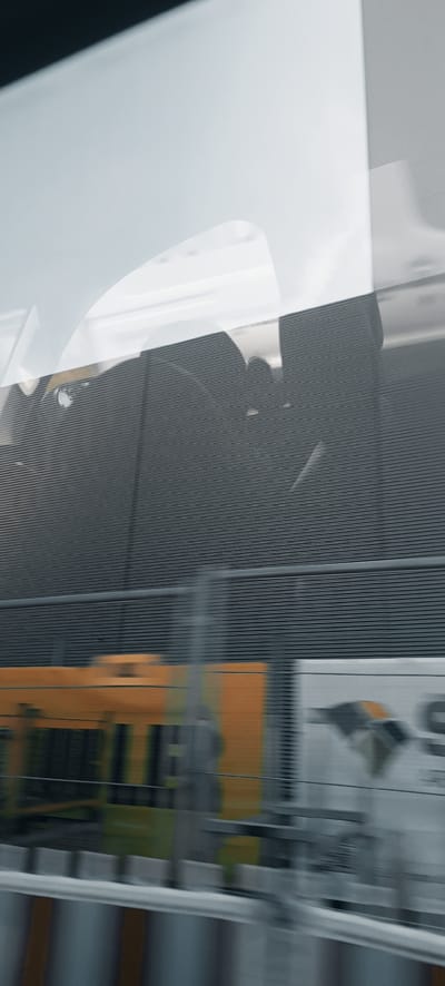 The image shows a motion-blurred view of a train traveling through an urban or industrial area. The foreground features a yellow and orange train car with some green accents, suggesting it might be a passenger or freight train. Behind the train, there is a dark gray wall with a horizontal ribbed texture, possibly a sound barrier or the side of a building. A lighter, reflective surface, perhaps a window or another building, is visible above the ribbed wall, showing abstract shapes and reflections. The overall impression is one of speed and transit, with the environment appearing somewhat indistinct due to the motion blur. The location is specified as Winterbach, Germany, implying this is a scene captured while the train was passing through or near the town.
