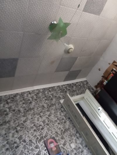 The image depicts an interior scene, likely a room, with a tiled ceiling and a patterned floor. A ceiling fan is visible, with a green decorative element above it. In the foreground, a partially opened white door frame is present, resting on the patterned floor. To the right, there are shelves with boxes and other items. The overall impression is one of a residential or storage space. The location is specified as Belovodskoe, Kyrgyzstan.