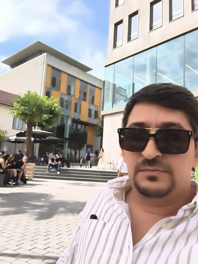 A selfie taken outdoors in Heilbronn, Germany, features a man in the foreground wearing sunglasses and a striped shirt. Behind him, a modern building with a glass facade and square windows dominates the background. To the left, a palm tree stands near a cafe with outdoor seating. Several people are visible in the mid-ground, some seated at tables and others walking. The ground is paved with interlocking bricks. The sky above is blue with scattered clouds.