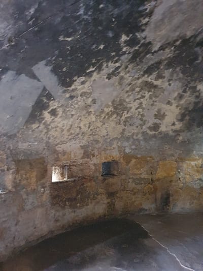 The image shows a close-up view of a textured, uneven, concrete wall in an indoor location. The wall is covered in a combination of grey, brown, and black. The concrete surface appears to be weathered and stained, possibly by water damage. There are two rectangular openings in the wall, suggesting the existence of a passage or ventilation system. It is impossible to determine the specific location or the use of the space, as there is no context or further information within the image. The image depicts a scene in Oran, Algeria (latitude: 35.701, longitude: -0.654). The location of the image is consistent with the provided coordinates. The scene contains no identifiable people or objects, only a textured wall with two openings.