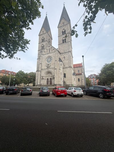 The image shows the exterior of the St. Andreas church in Würzburg, Germany, on an overcast day. The church features two prominent towers with spires, a large rose window above the main entrance, and stone construction. In front of the church, a row of cars is parked on a paved area. These cars include a black Audi, a gray Volkswagen Golf, a gray Fiat 500, a red Fiat 500, a white Seat Ibiza, and a black Kia Sportage. Trees with green foliage are visible on either side of the church and in the background. Residential buildings with red and white facades are also present in the background, with a German flag visible on one of them. The foreground is dominated by a wet asphalt road with a white line. There are no people visible in the image.