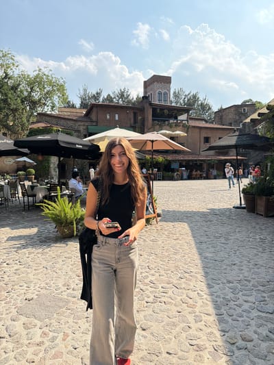 A young woman with long brown hair is smiling and holding a smartphone, seemingly taking a selfie. She is wearing a black T-shirt, light grey jeans, and red shoes. A black sweater is hanging from her shoulder.
The setting appears to be a pedestrian area in Domingo Arenas, Mexico, characterized by cobblestone pavement. There are buildings in the background with a mix of architectural styles, including a stone tower and structures with balconies. Outdoor seating for a restaurant or café is visible, with tables, chairs, and umbrellas. Several people are seated at the tables. The sky is partly cloudy.