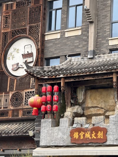 This image depicts a multi-story building facade in Qingyang District, China, showcasing a blend of traditional Chinese architectural and decorative elements with modern construction. The scene is devoid of people.

The left portion of the image features a traditional wooden facade composed of numerous intricate lattice panels with geometric and floral patterns, stained a reddish-brown. Centered on this facade, at mid-height, is a large circular white emblem with a raised relief depicting a traditional Chinese teapot, a teacup, and a branch with light green leaves.

To the right, a modern building constructed with dark grey bricks is visible, featuring rectangular windows with black frames and light-colored window sills. A vertical, light grey concrete column with a stepped, inverted-pyramid design extends upwards from just above a traditional roofline, reaching toward the upper windows of the brick building. A small metallic fixture, possibly a light or camera, is mounted on top of this concrete column.

Below the concrete column and the wooden facade, a traditional Chinese-style roof with dark grey, ridged tiles and ornate eaves juts out. The eaves are supported by intricately carved wooden pillars and beams. From beneath this roof, a cluster of red Chinese lanterns hangs. This cluster includes one larger spherical lantern with gold Chinese characters and a red tassel, and several smaller, cylindrical lanterns decorated with painted facial masks, arranged in vertical stacks.

Beneath the traditional roof, a facade designed to mimic natural rock formations is present, with varying shades of grey and beige, interspersed with small artificial greenery. At the bottom of the image, a grey brick wall, designed with crenellated battlements at its top, supports a rectangular wooden sign. The sign features raised gold Chinese characters that read "錦官城文創" (Jǐn Guān Chéng Wén Chuàng). Two small light fixtures are mounted on the grey brick wall, positioned to illuminate the wooden sign.