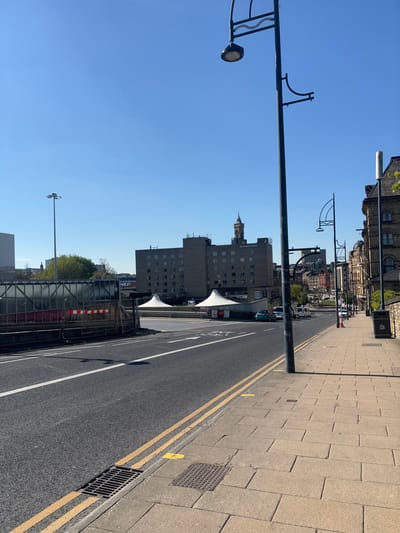 The image shows a street scene in Bradford, United Kingdom, taken at latitude 53.792 and longitude -1.751. The scene is captured from a pedestrian's perspective, looking down a road with two yellow lines marking the lanes. The road is paved with asphalt and is relatively empty with only a few vehicles in the distance. There are sidewalks on either side of the road, one of which the camera is positioned on. The sidewalk consists of grey brick pavers. There are a few street lights present, and the sky is a clear blue. In the background, there is a building with a tower on top, and other buildings can be seen in the distance. The scene is quiet and peaceful.