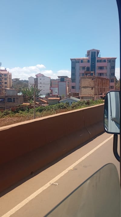 An urban landscape under a clear blue sky. Buildings of varying heights and designs populate the scene. In the foreground, a brown barrier wall runs parallel to a road with visible lane markings. Part of a vehicle, including a side mirror, is in the frame, suggesting the photo was taken from inside a car. Greenery mixes with rooftops, contrasting nature and human construction.