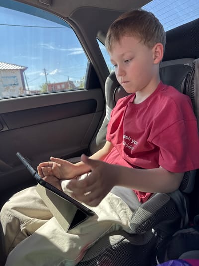 A young boy with fair hair is seated in a car, presumably in the back seat, as evidenced by the angle and the presence of a window to his left showing the outside. He is wearing a red t-shirt and khaki pants, and he is looking down at a tablet computer that he is holding with both hands. The surrounding environment is the interior of a car, with the car door and seat visible. The light suggests it is daytime, and through the car window, buildings and power lines are visible. It is not possible to confirm if this is Balezino, Russia from the image.