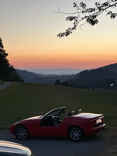 The image depicts a red convertible car parked on a gravel road at dusk. In the background, a person is walking on a paved path that winds up a grassy hill. Beyond the hill, layers of dark, silhouetted mountains are visible against a gradient sky of orange, pink, and pale blue, characteristic of a sunset. A branch with leaves hangs from the top right corner of the frame. The scene is likely set in a rural or suburban area outside of St. Peter, Germany, given the mountainous landscape and the presence of a paved walking path. The car is the primary subject in the foreground, with the natural landscape and the solitary figure in the background creating a serene and picturesque atmosphere.