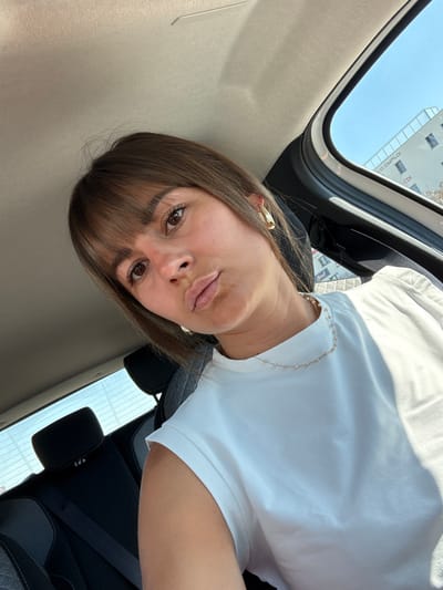 A woman is photographed inside a car, likely in Andrézieux-Bouthéon, France, based on the partial view of a building sign reflecting "CDI". She has brown hair with bangs and is wearing a white t-shirt and a gold necklace with beads. She is looking directly at the camera. The photo is taken at an angle, showing the car's interior including the roof, seat, and seatbelt. The outside sky is visible through the sunroof.