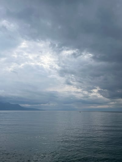 The image depicts a wide view of Lake Geneva from the city of Vevey, Switzerland, under an overcast sky. The foreground and midground consist of the calm, gray-blue water of the lake, showing subtle surface ripples. In the distant left, a dark, undulating silhouette of mountains forms the opposing shoreline. A single, small, dark vessel, likely a boat, is visible on the distant water surface towards the mid-right. The upper half of the frame is filled with dense, multi-layered gray clouds, with brighter, diffused light visible through thinner cloud sections, particularly in the upper left quadrant. There are no individuals, discernible actions, or interactions present in the scene. The environment is characterized by diffused natural light and a tranquil, overcast atmosphere.