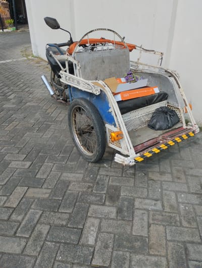 A weathered, three-wheeled motorized vehicle, possibly a becak or auto-rickshaw, is parked on a gray paver stone surface. The vehicle features a blue body with a white metal frame. In the rear cargo area, there is a black trash bag, two orange boxes stacked, and a small bunch of purple flowers. The vehicle has a single visible wheel with spokes. The background consists of a white wall and a section of a building with a doorway. The scene is set in Tanjungsari, Indonesia.