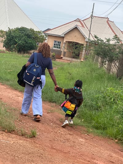 A woman with braided hair wearing a dark blue t-shirt and light blue pants walks hand-in-hand with a child wearing a brown jacket and a multicolored backpack. They are walking on a dirt path lined with tall green grass. In the background, there are two houses with peaked roofs and overcast skies. The woman is carrying a black bag over her left shoulder, and the child's backpack is strapped to their shoulders. The overall scene depicts a casual outdoor walk in a residential area.