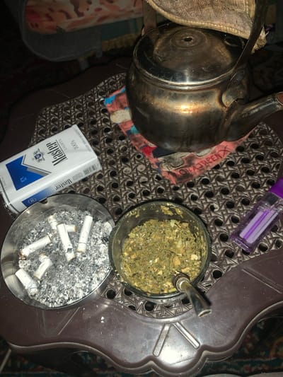 A cluttered tabletop scene features a silver teapot and a glass containing green, leafy material with a metal straw. Next to this is an ashtray filled with cigarette butts and ash. A pack of "Master" cigarettes and a purple lighter are also visible. The tabletop has a woven pattern with some dark, solid sections. The overall impression is one of smoking paraphernalia and a casual setting.