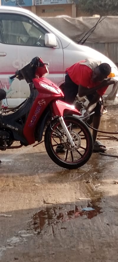 This image depicts an outdoor scene in Kabuga, Nigeria, likely at a vehicle washing facility during the daytime. The central subject is a man, dressed in a red t-shirt and dark shorts or trousers, actively washing a red motorcycle. He is bent over the front section of the motorcycle, holding a nozzle or hose to direct water onto the vehicle.

The motorcycle is a red scooter-style model with black accents, including its seat and some lower body panels. Distinct text on its side reads "CAR-GO," accompanied by a stylized horse logo, and "CAR110-80" near the engine. The motorcycle features black wheels with striking red rims and a disc brake system on the front wheel. The ground beneath and around the vehicles is wet and muddy, consistent with a washing operation.

In the background, a white car, resembling an SUV or pickup truck, is parked. Its side mirror is visible, and its windshield wipers are raised, suggesting it might also be in the process of being washed or recently completed. Faint, partial text, possibly "Ch...", can be seen on the side of the white car. Further in the background, a sign with the clear text "Steel Work" is visible, hinting at a nearby business. The natural lighting suggests it is daytime. The man appears focused on his work.