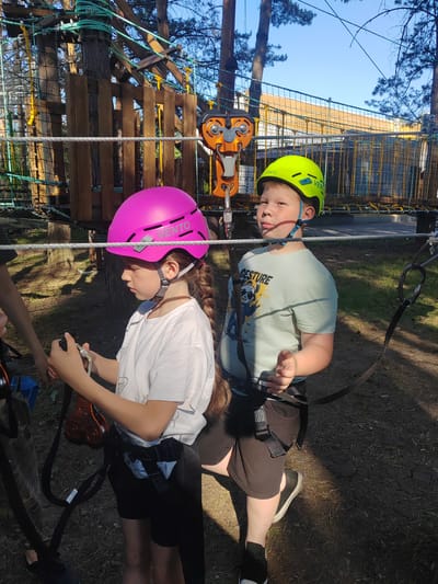 Two children, a girl and a boy, are participating in a ropes course activity. The girl, on the left, wears a pink helmet and a white t-shirt. She has a long braid and is holding onto a carabiner and harness. The boy, on the right, sports a bright yellow helmet and a light blue t-shirt with a graphic print. He is also wearing a harness. They are both secured by safety lines.

The setting is an outdoor adventure park, likely a ropes course constructed among trees. The structures visible include wooden platforms, netting, and thick ropes, with a bright orange zipline pulley system present. The ground is covered in dirt and dry leaves, suggesting a sunny and possibly dry day. The sunlight casts distinct shadows, indicating it is likely midday. The overall mood appears to be one of focused engagement with the activity, with the boy looking slightly to the side with a neutral expression and the girl concentrating on her equipment. The scene is set in a natural environment with trees providing shade and structure. The location is identified as Berezovo, Russia. Visible text on the boy's helmet reads "VENTO" multiple times.
