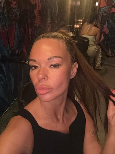 A woman with her hair in a ponytail and wearing a black tank top is taking a selfie. She is in the foreground, with her face turned towards the camera. Her lips are full and slightly parted, and she has noticeable makeup, including eyeliner and blush. A small nose ring is visible on her right nostril. Her right hand is touching her hair, and her left arm is partially visible, revealing a tattoo on her upper arm. In the background, another person is seated, wearing light-colored clothing. The environment appears to be an indoor setting with patterned wallpaper and dim lighting, suggesting a bar or restaurant. There is a large black bowl-like object to the left of the woman in the foreground, and another darker object to her right, possibly furniture. The overall impression is a casual, possibly social setting. The location is specified as Ban Bang Thao, Thailand.