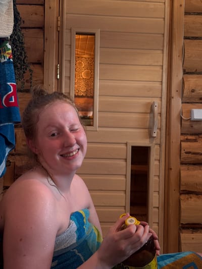 A young woman with red hair pulled up into a bun is sitting in a sauna, wrapped in a blue and yellow towel. She is winking and smiling broadly at the camera, with sweat visible on her skin. She is holding a glass bottle with a yellow cap, seemingly about to open it. The sauna is made of light-colored wood paneling with a small window showing a patterned interior. To the left, a blue towel with red and white lettering hangs on the wall. To the right of the sauna door, a wooden handle is attached to the paneling, and a white light switch is mounted on a rough-hewn log wall. The overall environment suggests a rustic, wooden sauna, possibly located in or around the city of Gubakha, Russia, given the context provided.