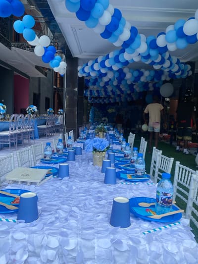 The image shows a party table set up outdoors. The table is covered in a white tablecloth with blue petal decorations and features a long line of blue plastic cups and blue plastic plates. The party is decorated with blue and white balloons strung along the ceiling. Several people are in the background, including a man in a white shirt and blue shorts, who is walking. There are white plastic chairs on either side of the table and a green lawn in the background. Bottles of water with blue labels are on the table, and there is a small floral arrangement in a gold vase. There is a small plastic plate with a white paper on top in front of a plastic cup. There are wooden spoons and a paper straw with blue and white stripes on each place setting. This is a festive outdoor party setting, likely at a home in Limbe, Cameroon.