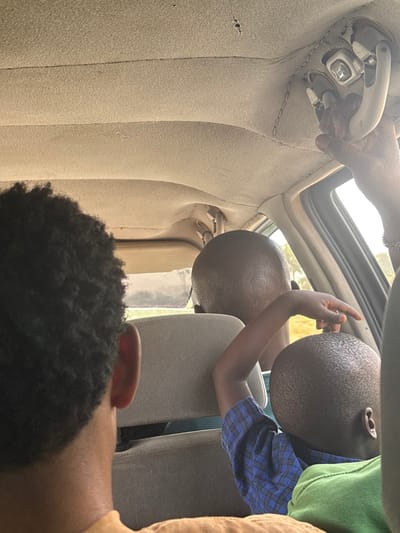 The image depicts the interior of a vehicle in Marke, Nigeria, during daylight hours. From the perspective of a rear passenger, the scene shows at least four individuals. In the immediate foreground, the back of a person's head with dark, curly hair and a visible ear is seen, wearing a light brown garment. Directly in front of this individual, partially obscured by a grey seat headrest, is the back of an adult's shaved head. To the right, a child with a shaved head, wearing a blue plaid shirt and a green garment, is seated. This child's left arm is raised, with their hand gently touching the crown of the shaved head in front of them. In the upper right quadrant, a dark-skinned hand adorned with a silver bracelet grips an overhead grab handle, which appears to be part of a damaged or exposed console on the beige, slightly sagging vehicle headliner. Light filters in from windows on the right side of the vehicle, revealing a blurry, sunlit outdoor environment consistent with natural terrain.