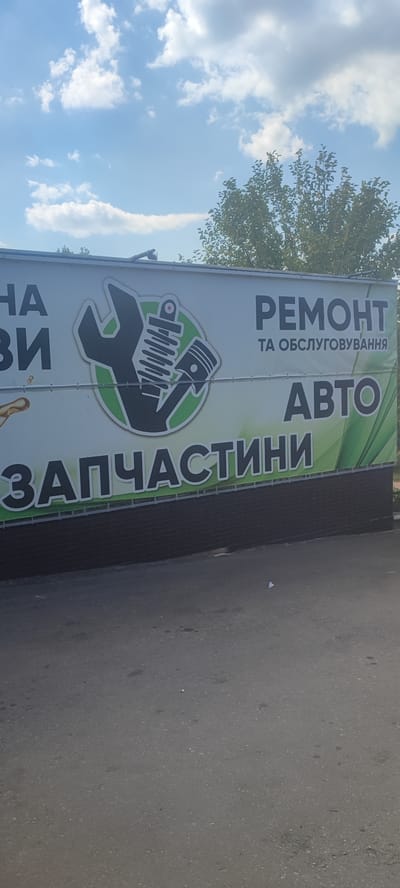 A wide, horizontal banner is displayed on the side of a building under a bright, partly cloudy sky. The banner features large, bold text and a graphic logo.
On the left side of the banner, partially visible text in black reads "НА" and "ВИ". To the right of this text is a logo consisting of a large black wrench and a car shock absorber with a piston, all enclosed within a green circle.
The right side of the banner prominently displays the word "РЕМОНТ" in large black letters, with smaller text below it reading "ТА ОБСЛУГОВУВАННЯ". Further down, in even larger lettering, is the word "АВТО".
The bottom section of the banner features the word "ЗАПЧАСТИНИ" in large black letters against a green background that transitions to a darker green at the bottom. The banner is attached to a dark brick-like structure, and the ground in front of it is asphalt.
In the background, green trees are visible against the blue sky dotted with white clouds. The lighting suggests it is daytime. There are no people or explicit activities depicted, but the text clearly indicates the services offered are auto repair, maintenance, and parts, likely at a business in Svitlovodsk, Ukraine.