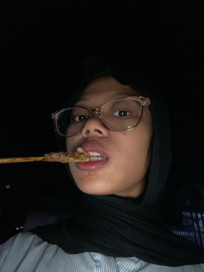 A young woman wearing glasses and a black hijab is eating satay from a skewer in Tanggul, Indonesia. She is looking at the camera and her mouth is partially open as she bites into the food. The background is dark, suggesting it is nighttime or indoors with low lighting. She is wearing a light blue and white striped shirt.