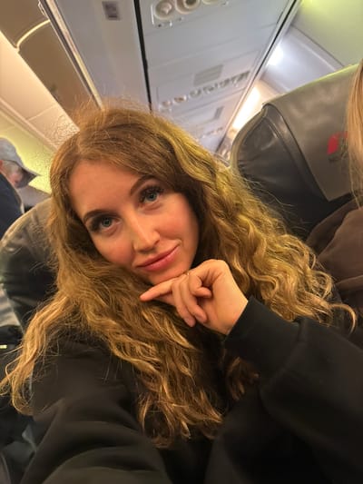 A woman with long, curly blonde hair and blue eyes rests her head on her hand inside an airplane cabin. She wears a black hooded sweatshirt. The interior of the plane is visible, including the overhead compartments, seats, and control panels. This image was taken in an airplane that just took off or landed in the city of Bolshoye Savino, Russia.